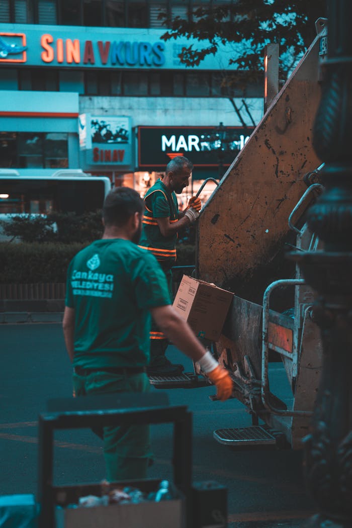 Two workers operate a garbage truck in a city street at night, wearing uniforms.